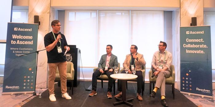 A speaker stands with a microphone next to a seated panel of three professionals during a session at ascend grc day in toronto. Two branded signs on either side read “welcome to ascend” and “connect. Collaborate. Innovate. "