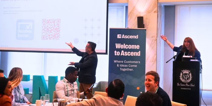 Two presenters enthusiastically gesture toward a large screen during a session at ascend grc day in toronto, while attendees seated at round tables look on and laugh. A nearby banner reads “welcome to ascend – where customers & ideas come together. ”