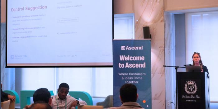 A speaker stands at a podium presenting at ascend grc day in toronto, with a projected slide titled “control suggestion” visible behind her. Attendees are seated at tables, and a nearby banner reads “welcome to ascend – where customers & ideas come together. ”