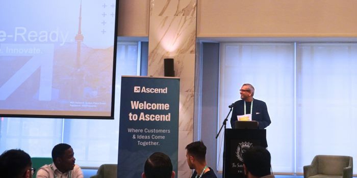 Resolver president, kam rawal, stands at a podium addressing attendees at ascend grc day in toronto. A large banner beside him reads “welcome to ascend – where customers & ideas come together,” while a screen in the background displays a slide with the cn tower faintly visible. Seated audience members listen attentively.