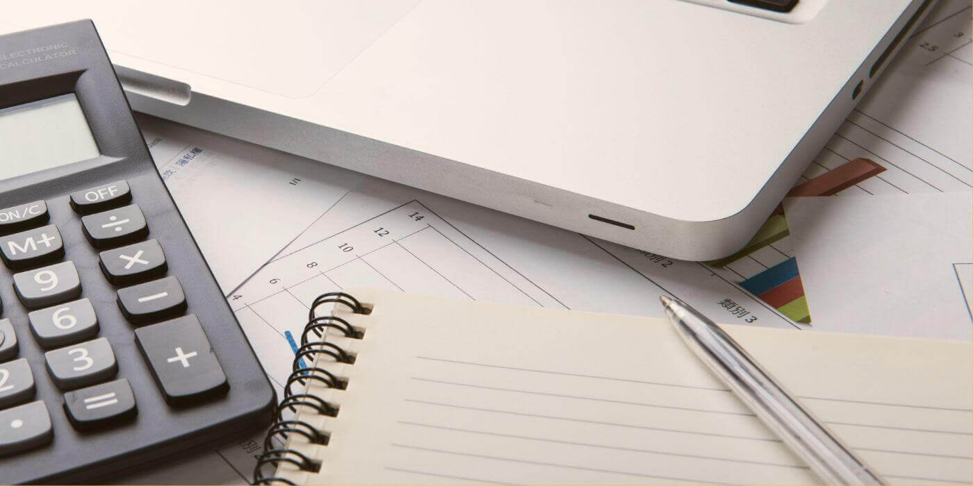 Close-up of a desk setup featuring a calculator, open notebook with a pen, and a laptop, all placed on top of financial documents and charts, representing tools and strategies used in detecting systemic fraud.