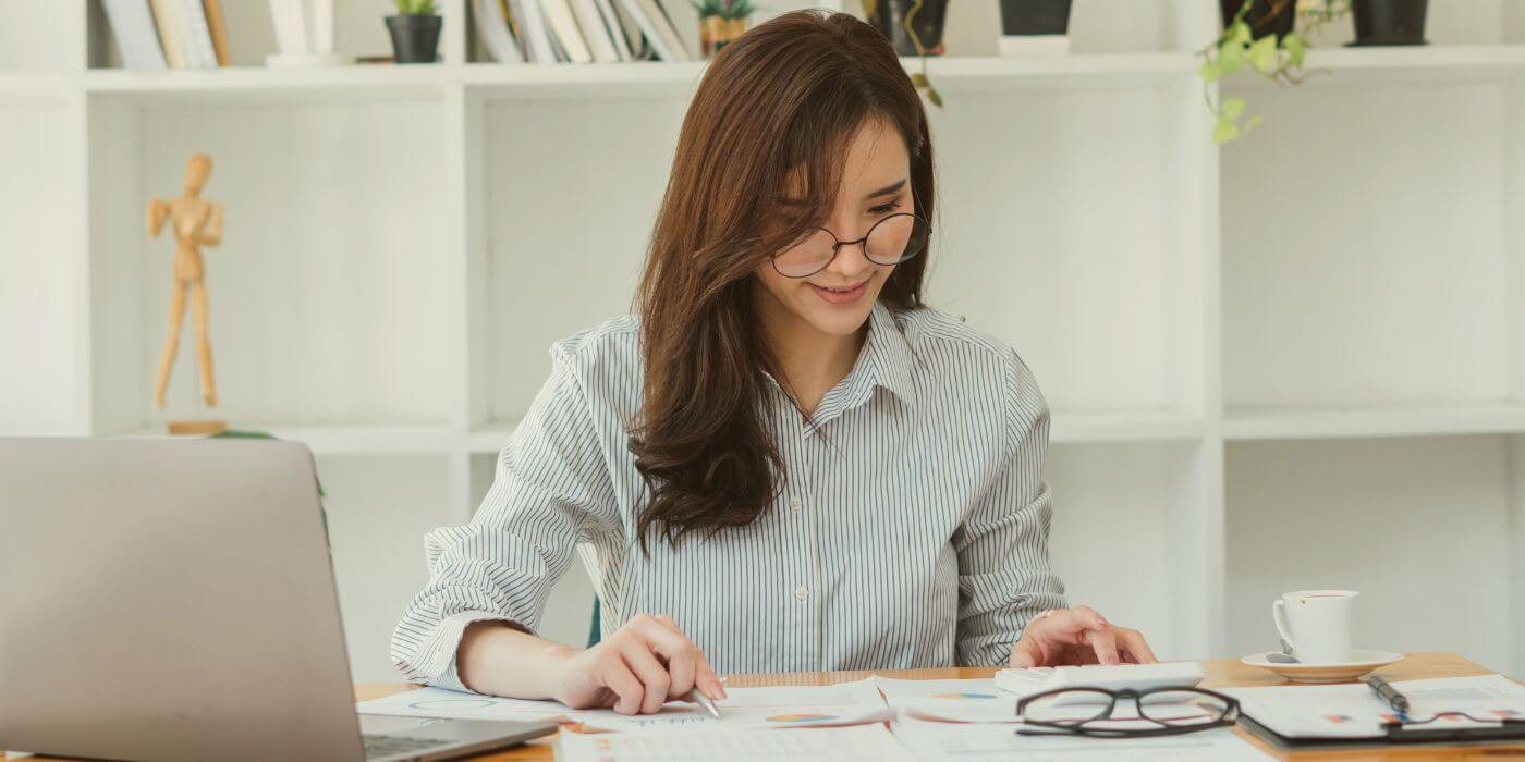 Business professional reviewing internal controls at her desk, focusing on charts and documents while working on a laptop.
