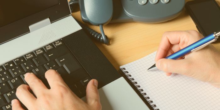 One hand typing on laptop, another holding a pen to write in a notebook representing the benefits of risk management software