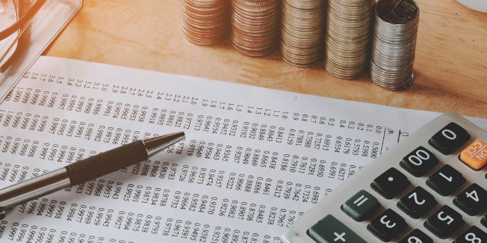 Pen on spreadsheet with numbers in foreground with calculator and stacks of coins in the background representing regulatory compliance for banks