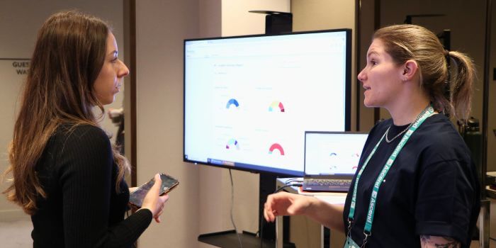 Ascend 2025: what security teams are really solving for 4 Two women in conversation at a tech demo during a conference, with data dashboards displayed on a monitor and laptop screen in the background.