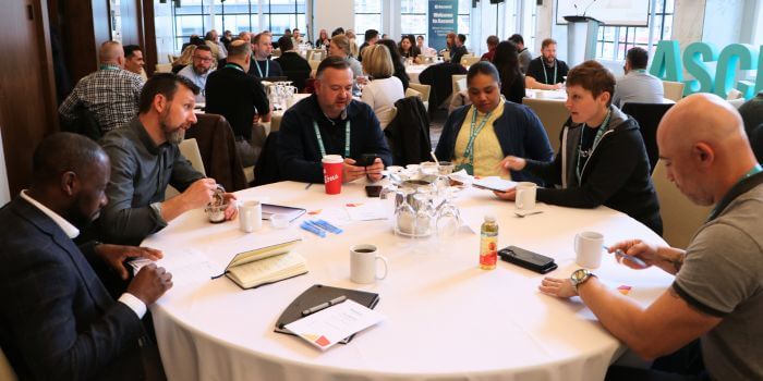 Ascend 2025: what security teams are really solving for 2 Attendees seated around a round table engaged in discussion during a conference session, with notebooks, drinks, and event lanyards visible; the large teal "ascend" letters can be seen in the background.