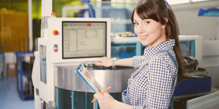 A female technician with brown hair, wearing a checkered shirt, smiling at the camera while holding a clipboard and pen, standing in front of a manufacturing machine with a digital control panel representing manufacturing access control