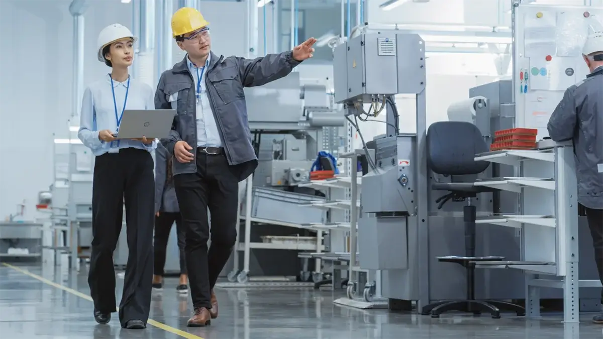 Female engineer with a laptop and male supervisor wearing safety helmets discuss over the operational aspects beside industrial machinery, highlighting active use of manufacturing plant security software from Resolver on the facility floor.